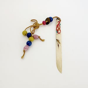 May include: A decorative letter opener with a bone-colored blade and a beaded tassel. The tassel features yellow, blue, and pink beads, and is attached to the opener with a knotted cord. The blade has painted details.