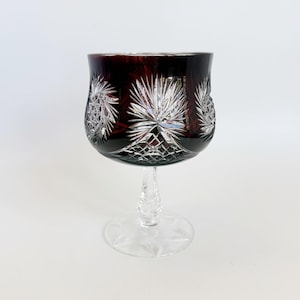 May include: A ruby red crystal wine glass with intricate starburst and geometric cut patterns. The stem is clear and features a twisted design, and the base is a clear, star-shaped design. The glass is likely used for serving wine.