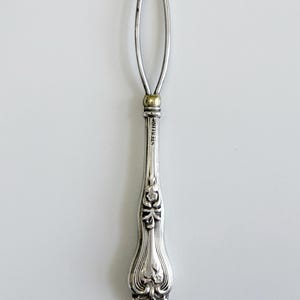 May include: A silver button hook with a long, thin, oval-shaped wire loop at the top and a decorative, ornate handle. The handle features embossed floral patterns and the word STERLING is clearly stamped vertically along the metal shaft.