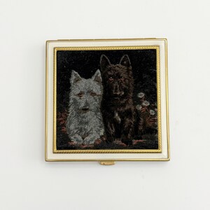 May include: A square compact with a gold-colored frame and a white border. The center features a tapestry-like image of two dogs, one gray and one black, against a dark background. The compact has a small clasp.