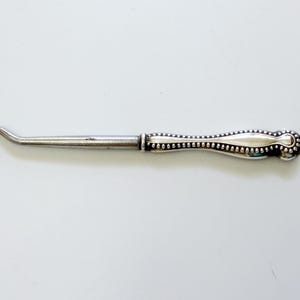 May include: A vintage silver crochet hook with a curved end and a decorative handle. The handle features a beaded design and a rounded end. The hook is designed for crafting and textile work.