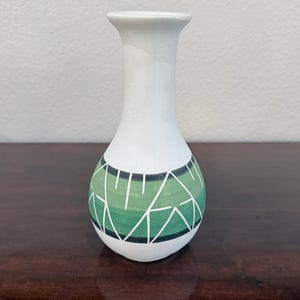 May include: A white ceramic vase with a narrow neck and a bulbous base. The lower section features a horizontal band of varying shades of green, decorated with a series of white geometric lines forming triangles and angular shapes throughout.