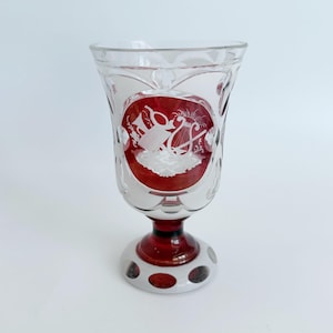 May include: A clear and red glass goblet featuring a circular medallion with an etched white design of a lyre and musical instruments. The stem and base are red with white circular accents, and the bowl has a scalloped rim and cut glass details.