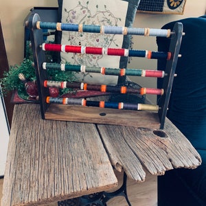 Puede incluir: Un estante de madera para hilos con cinco estantes que contienen carretes de hilo de colores. El estante es de color marrón oscuro y tiene un aspecto rústico. El hilo está en varios tonos de azul, rojo, verde y naranja.