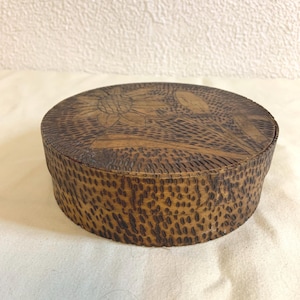 Könnte beinhalten: Eine runde Holzkiste mit Deckel. Der Deckel zeigt ein geschnitztes Blumenmuster. Die Seiten der Kiste weisen ein strukturiertes, gepunktetes Muster auf. Das Holz hat eine warme, braune Farbe. Die Kiste steht auf einer weißen Oberfläche.