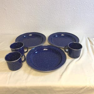 Puede incluir: Un juego de vajilla de camping de esmalte azul moteado. El juego incluye tres platos y dos tazas. Los platos y las tazas tienen un borde plateado. La vajilla está dispuesta sobre un mantel blanco.