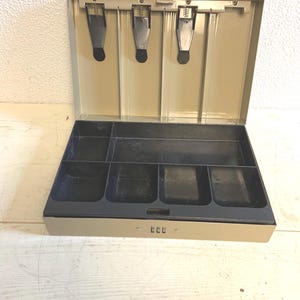 Puede incluir: Una caja registradora metálica beige abierta, con interior negro. Cuenta con tres compartimentos para billetes con resorte y varios compartimentos para monedas. Se ve un candado de combinación.