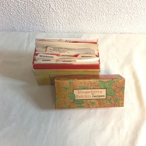 May include: Two vintage recipe boxes. One box is open, filled with recipe cards labeled "Beverages", "Soups", "Salads", and "Desserts". The other box is closed, with a design of green leaves and the text "Strawberry Patches recipes".