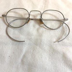 May include: Vintage round eyeglasses with a silver metal frame. The lenses are clear, and the earpieces are detached. The glasses have a bridge and nose pads. The frame shows signs of wear, suggesting age.