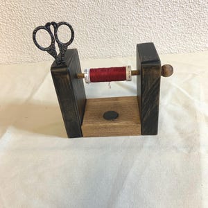 Puede incluir: Un portahilos de madera con un carrete de hilo rojo y un par de tijeras pequeñas y ornamentadas. El soporte está hecho de madera teñida oscura y madera natural, con un pequeño imán en la base. Un artículo de artesanía.