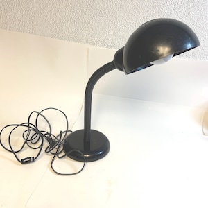 May include: A black desk lamp featuring a dome-shaped shade, a flexible gooseneck arm, and a circular base. The lamp includes a black power cord with an inline switch, and a white light bulb is visible inside the shade of the lamp.