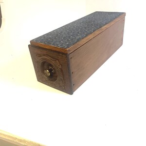 May include: A rectangular wooden box with a dark brown finish and a hinged lid. The top surface is covered in a dark blue fabric with a small white star pattern. The front panel features an ornate carved circular design with a small brass knob.