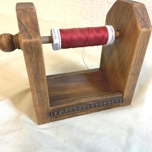 Peut inclure: Un porte-fil en bois avec une bobine de fil rouge. Le fil est étiqueté avec le texte "yds. col. 601". Le support est en bois teinté et possède un bord décoratif perlé.