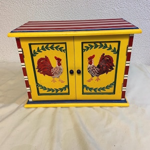 May include: A yellow wooden cabinet with two doors, painted with red and black roosters and green leaves. The top of the cabinet has red and white stripes.