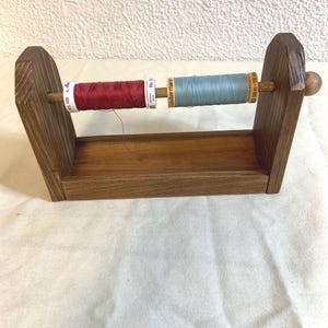 May include: A wooden thread spool holder with two spools of thread. One spool is red, the other is blue. The holder is made of dark brown wood and has a small shelf at the bottom.