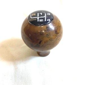 以下が含まれることがあります： 茶色の大理石模様のヴィンテージカーのギアシフトノブ。上部には、ギアの位置を示す白文字と数字が書かれた黒いプレートがあり、「R」、「1」、「2」、「3」、「4」が含まれています。「PRESS」という文字も表示されています。