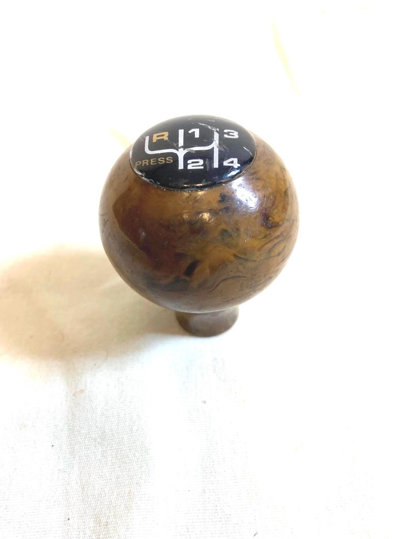 以下が含まれることがあります： 茶色の大理石模様のヴィンテージカーのギアシフトノブ。上部には、ギアの位置を示す白文字と数字が書かれた黒いプレートがあり、「R」、「1」、「2」、「3」、「4」が含まれています。「PRESS」という文字も表示されています。