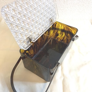 May include: A vintage-style, rectangular handbag with a clear, patterned lid and a dark, tortoiseshell-patterned body. The bag has a matching handle and is open, revealing the interior.