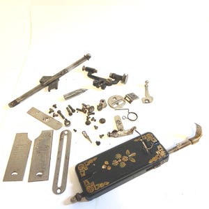 Könnte beinhalten: Eine Sammlung von Nähmaschinenteilen, darunter eine schwarze, rechteckige Metallplatte mit goldenen Blumenmustern, verschiedene Stahlschrauben, Metallplatten, eine lange Stange und mechanische Komponenten, die auf einer weißen Fläche liegen.