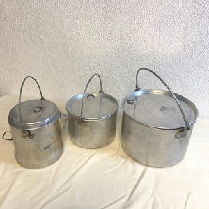 Op de afbeelding: Drie vintage aluminium kampeerkannen met deksels en draadgrepen. De potten hebben verschillende maten, met een waterkoker links en twee ronde potten. De potten staan op een wit oppervlak.