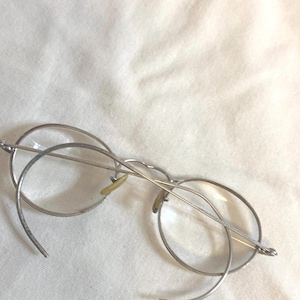 May include: Silver-framed eyeglasses with round lenses. The frames have a unique, curved design extending beyond the lenses. The nose pads are a light yellow color. The glasses are set against a white background.