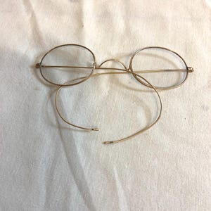 Könnte beinhalten: Antike goldfarbene Brille mit runden Gläsern. Die Brille hat einen filigranen Drahtrahmen und gebogene Bügel. Der Rahmen ist auf einer Seite gebrochen. Die Brille liegt auf einer weißen Oberfläche.