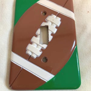 Puede incluir: Una tapa de interruptor de luz en forma de balón de fútbol marrón y blanco con fondo verde.