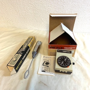 Puede incluir: Un surtido de electrodomésticos de cocina antiguos, que incluyen una batidora a pilas y un temporizador automático de lámpara de 24 horas. La batidora está en su caja original y el temporizador tiene una esfera de reloj en blanco y negro. Los artículos están sobre una superficie blanca.