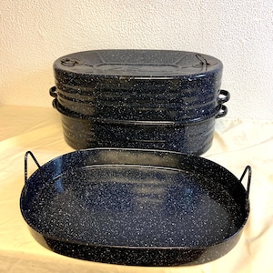 May include: A stack of dark blue enamelware roasting pans with lids, and a single oval pan in the foreground. The pans have a speckled white pattern and black handles. The pans are likely vintage or retro.