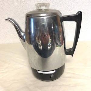 Puede incluir: Una cafetera eléctrica de percolador en cromo pulido con mango y base de plástico negro. La unidad cuenta con un pomo de cristal transparente en la tapa y un pequeño interruptor deslizante negro con el texto MILD y STRONG impreso.