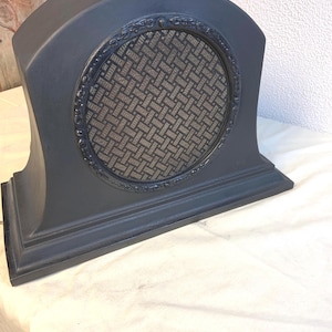 May include: A dark grey mantel clock with a decorative circular design. The clock features a patterned mesh screen within a detailed frame. The clock has a curved top and a rectangular base, showcasing a classic design.