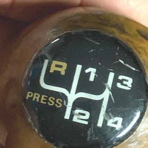 以下が含まれることがあります： 黒い文字盤が付いた木製のギアシフトノブ。「R」、「1」、「2」、「3」、「4」のギア位置を示す白文字と線があります。「PRESS」の文字も表示されています。