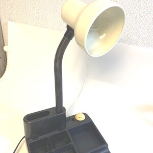 May include: A desk lamp featuring a cream-colored adjustable head attached to a flexible black neck. The base is a black plastic organizer with multiple compartments for storage and a small round cream-colored power button on the top surface.