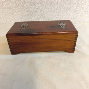 May include: A wooden box with two silver metal accents on the top. The box is made of dark wood with a natural grain pattern.