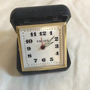 May include: A vintage travel alarm clock with a square face and gold-colored trim. The clock face is white with black numerals and hands. The clock is housed in a black, hinged case. The word "EQUITY" is printed on the clock face.