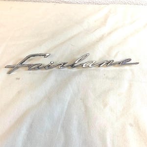 May include: A chrome "Fairlane" emblem in a cursive script. This car badge is likely from a classic vehicle. The emblem has a reflective surface and is set against a white background. The emblem is a vintage car part.