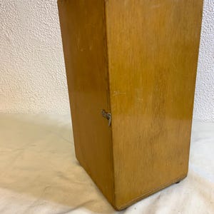 May include: A vintage wooden box with a hinged lid and a metal clasp. The box is rectangular with a light brown finish and visible wood grain. The box is likely used for storage.