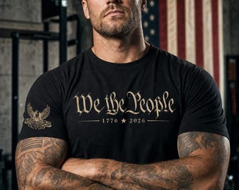 May include: A black short-sleeve t-shirt featuring the text We the People in a gold-toned font across the chest, with the years 1776 and 2026 separated by a star. A small gold eagle emblem is printed on the right sleeve of the garment.