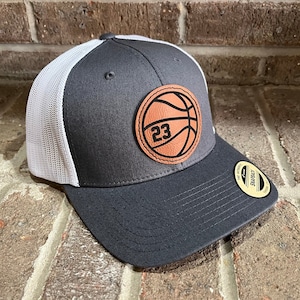 May include: A gray and white trucker hat with a brown leather basketball patch featuring the number 23.