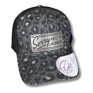 May include: A black and gray leopard print baseball cap with a black mesh back. The cap features the text "Sorry About My Husband" on a silver rectangular patch. A white and purple sticker is attached to the brim.