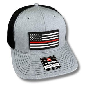 May include: Gray trucker hat with a black mesh back and a curved brim. The hat features a patch of the American flag with a red stripe. The Richardson brand label is visible on the front.