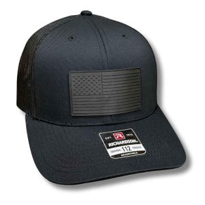 May include: Black trucker hat with a mesh back and a curved brim. The front features a black American flag patch. The hat includes a Richardson 112 tag, indicating an adjustable fit.