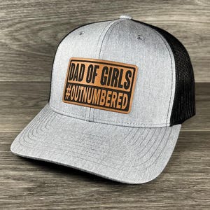 May include: A gray and black trucker hat with a brown leather patch that reads "Dad of Girls #Outnumbered".