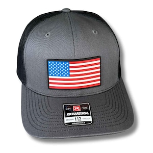 May include: Gray trucker hat with a black mesh back and a patch featuring the American flag. The hat has a curved brim and an adjustable strap. The Richardson brand label is visible.
