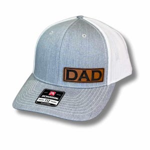 May include: A gray and white trucker hat with a curved brim. The hat features a brown leather patch with the word "DAD" in bold, black letters. The hat has a mesh back and an adjustable strap.