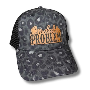 May include: A black and white leopard print trucker hat with a brown leather patch that says "Somebody's Problem".
