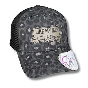 May include: Black and white leopard print trucker hat with a silver patch that reads "I Like My Men A Little Bit Younger".