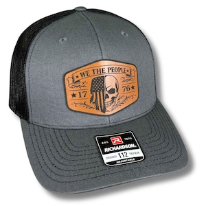 May include: Gray trucker hat with a black mesh back. The front features a leather patch with the text "WE THE PEOPLE", a skull, and an American flag design. The hat has a curved brim and an adjustable strap.