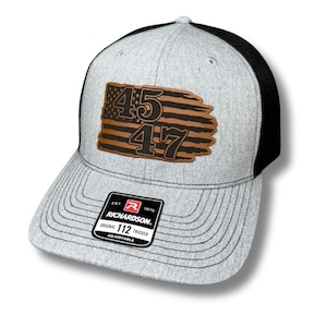 May include: A gray and black trucker hat with a brown leather patch featuring an American flag with the numbers 45 and 47. The hat is adjustable and has the Richardson 112 logo on the back.