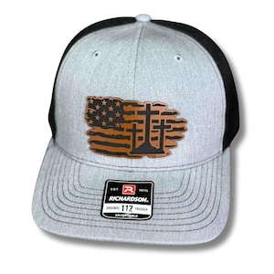 May include: A gray and black trucker hat with a leather patch featuring an American flag design with three crosses. The hat has a curved brim and an adjustable strap. The Richardson brand label is visible.